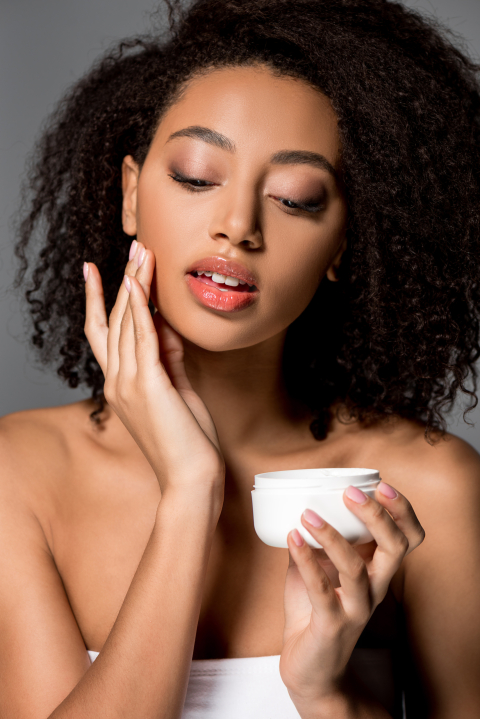 tender young african american woman applying face cream, isolated on grey