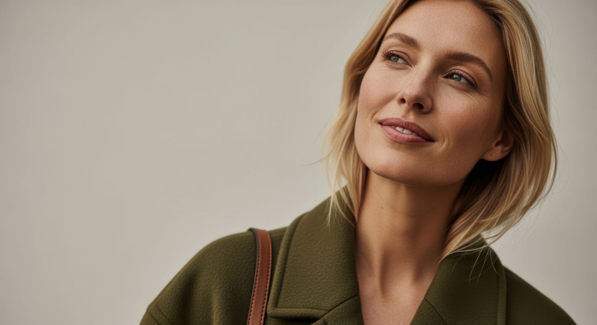 a young caucasian woman with short blond hair smiles softly. she wears a green coat and has a brown shoulder bag. the background is neutral and minimalistic.